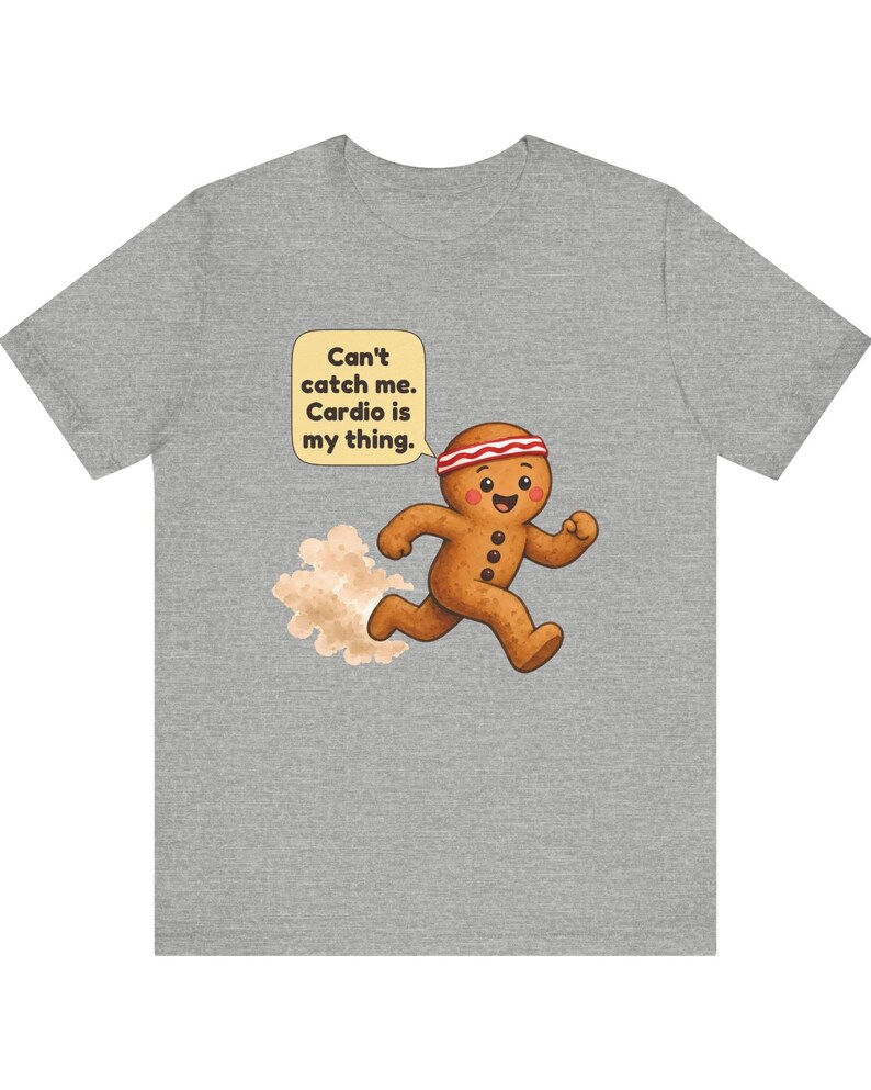 May include: A heather grey t-shirt featuring a cartoon gingerbread person running with a red and white headband. A speech bubble above the character contains the text: Can't catch me. Cardio is my thing. A small cloud of dust follows the figure.
