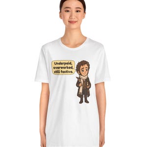 May include: A white t-shirt featuring a cartoon illustration of a person in a brown coat and white scarf holding a quill and candle. A yellow speech bubble next to the character contains the text: Underpaid, overworked, still festive.