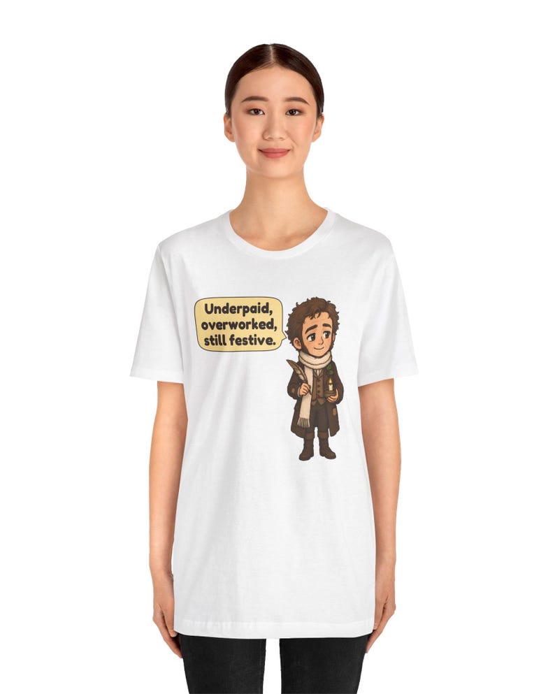 May include: A white t-shirt featuring a cartoon illustration of a person in a brown coat and white scarf holding a quill and candle. A yellow speech bubble next to the character contains the text: Underpaid, overworked, still festive.