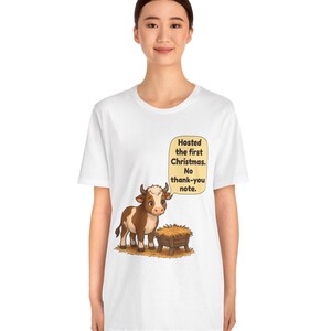 以下が含まれることがあります： 白い半袖Tシャツ。干し草が入った木製の飼い葉桶の横に立つ、茶色と白の漫画風の牛が描かれています。牛の上の吹き出しには「Hosted the first Christmas. No thank-you note.」というテキストが書かれています。