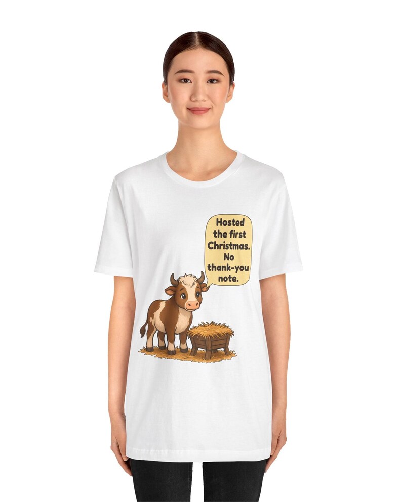 以下が含まれることがあります： 白い半袖Tシャツ。干し草が入った木製の飼い葉桶の横に立つ、茶色と白の漫画風の牛が描かれています。牛の上の吹き出しには「Hosted the first Christmas. No thank-you note.」というテキストが書かれています。