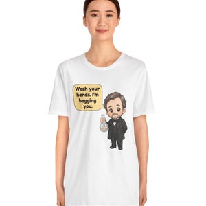 May include: A white t-shirt featuring a cartoon illustration of a man in a black suit holding a glass flask. A yellow speech bubble next to the character contains the black text: Wash your hands. I'm begging you.