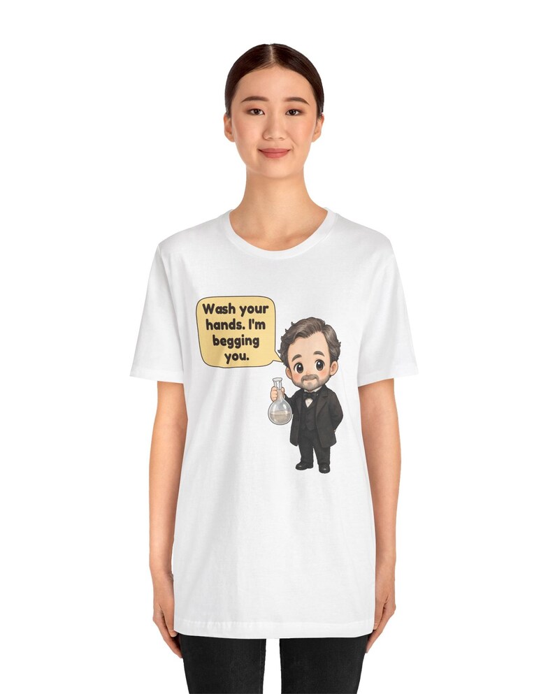 May include: A white t-shirt featuring a cartoon illustration of a man in a black suit holding a glass flask. A yellow speech bubble next to the character contains the black text: Wash your hands. I'm begging you.