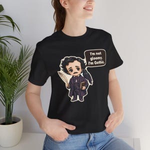 May include: A black short-sleeved t-shirt featuring a cartoon illustration of a man with dark curly hair, a moustache, and a dark coat, holding a quill and book with a raven on his shoulder. A speech bubble contains the text: I'm not gloomy. I'm Gothic.
