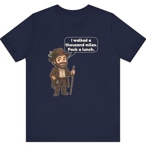 May include: A navy blue t-shirt featuring a cartoon illustration of a bearded hiker wearing a brown hat with a pine sprig, a brown coat, and boots, holding a wooden walking stick. A speech bubble contains the text: I walked a thousand miles. Pack a lunch.