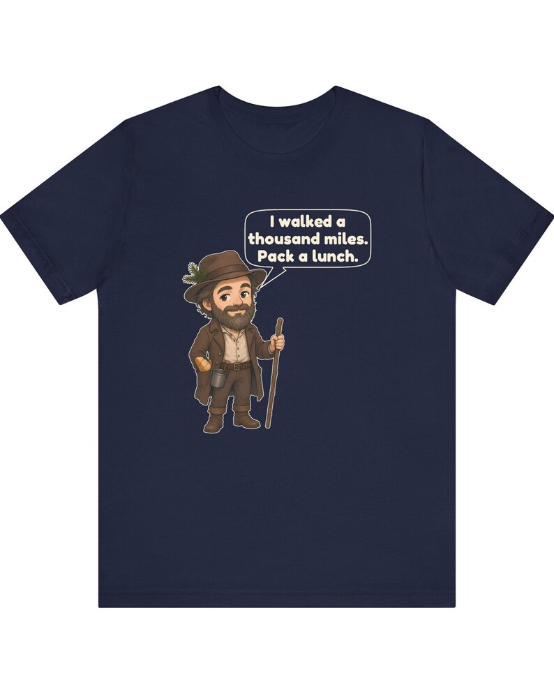 May include: A navy blue t-shirt featuring a cartoon illustration of a bearded hiker wearing a brown hat with a pine sprig, a brown coat, and boots, holding a wooden walking stick. A speech bubble contains the text: I walked a thousand miles. Pack a lunch.