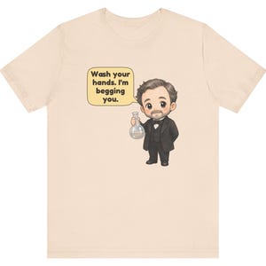 May include: A light beige t-shirt featuring a cartoon illustration of a man in a black suit and bowtie holding a glass flask. A speech bubble next to him contains the text: Wash your hands. I'm begging you. The design is centered on the chest.