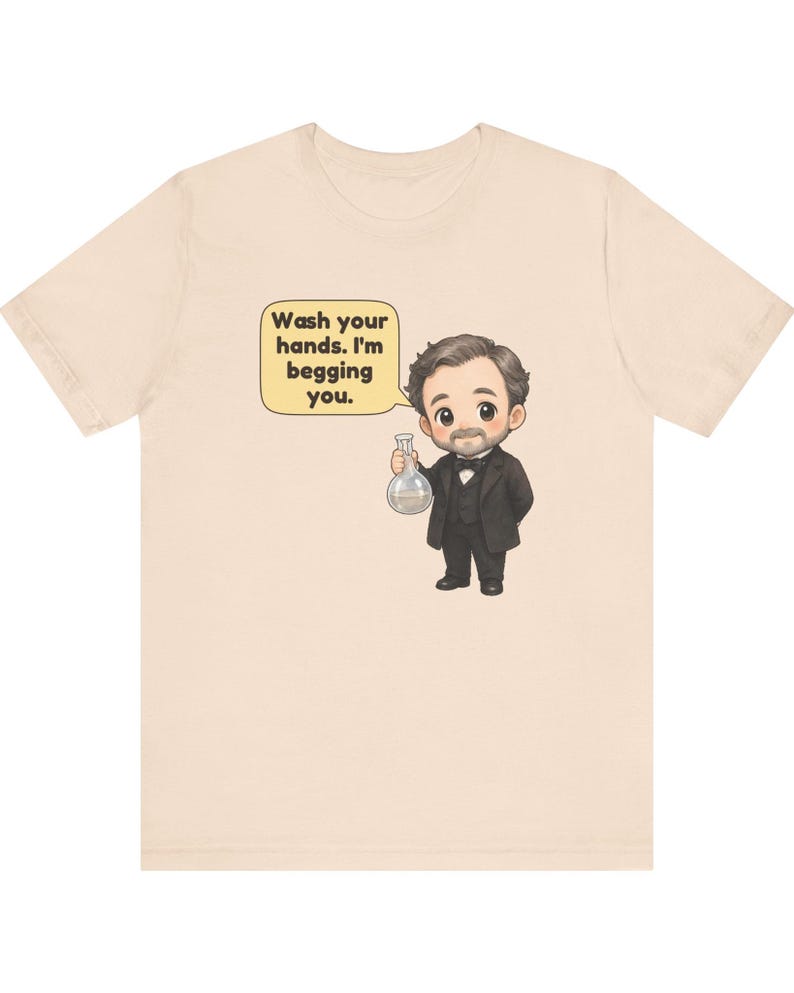 May include: A light beige t-shirt featuring a cartoon illustration of a man in a black suit and bowtie holding a glass flask. A speech bubble next to him contains the text: Wash your hands. I'm begging you. The design is centered on the chest.