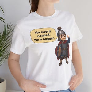 May include: A white t-shirt featuring a cartoon illustration of a warrior in chainmail armor, a red cape, and a helmet, holding a round shield. A yellow speech bubble contains the text: No sword needed. I'm a hugger.