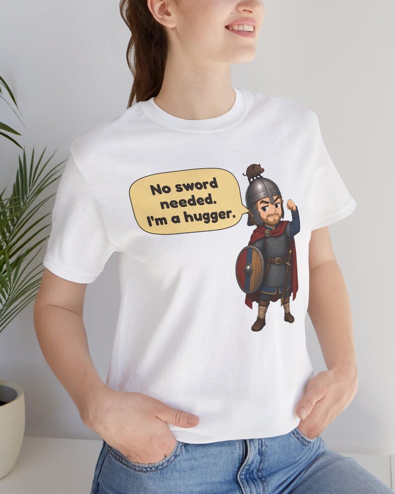 May include: A white t-shirt featuring a cartoon illustration of a warrior in chainmail armor, a red cape, and a helmet, holding a round shield. A yellow speech bubble contains the text: No sword needed. I'm a hugger.