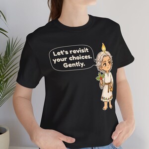 May include: A black t-shirt featuring a graphic of a light-haired figure with a candle flame on their head, holding holly. A speech bubble contains the text: Let's revisit your choices. Gently. The figure wears a white tunic with floral trim.