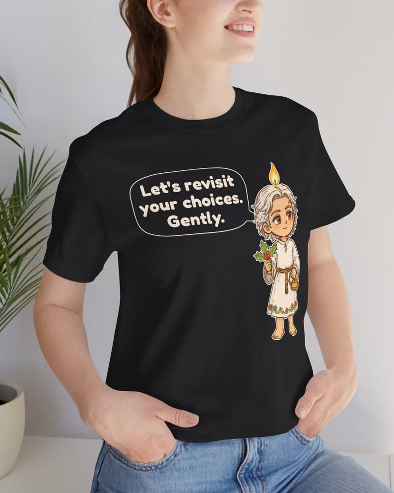 May include: A black t-shirt featuring a graphic of a light-haired figure with a candle flame on their head, holding holly. A speech bubble contains the text: Let's revisit your choices. Gently. The figure wears a white tunic with floral trim.