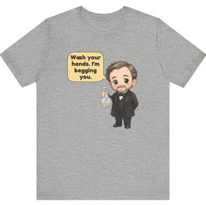 May include: A heather gray t-shirt featuring a cartoon illustration of a man in a black suit and bowtie holding a glass flask. A speech bubble next to him contains the text: Wash your hands. I'm begging you.
