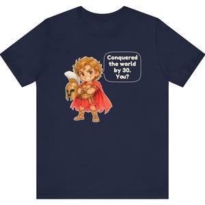 May include: A navy blue short-sleeve t-shirt featuring a cartoon illustration of a warrior in golden armor and a red cape holding a helmet. A speech bubble next to the character contains the text: Conquered the world by 30. You?