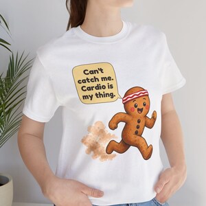 May include: A white t-shirt featuring a cartoon gingerbread person running with a dust cloud behind it. The gingerbread person wears a red and white headband. A yellow speech bubble contains the text: Can't catch me. Cardio is my thing.