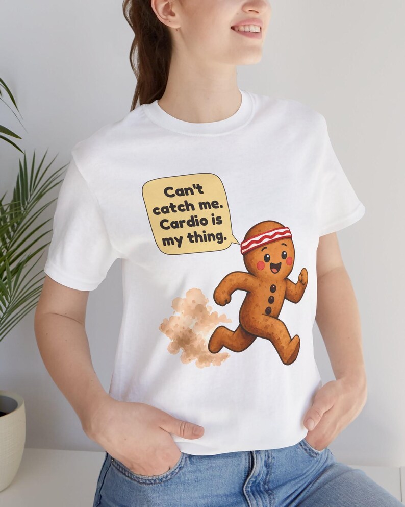May include: A white t-shirt featuring a cartoon gingerbread person running with a dust cloud behind it. The gingerbread person wears a red and white headband. A yellow speech bubble contains the text: Can't catch me. Cardio is my thing.