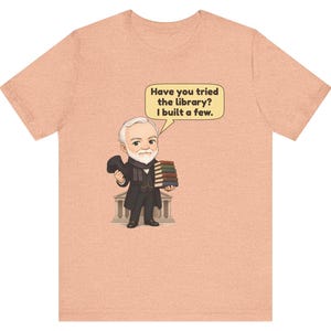 May include: A peach-colored t-shirt featuring a cartoon illustration of a bearded man in a suit holding a stack of books and a hat. A speech bubble above him contains the text: Have you tried the library? I built a few.