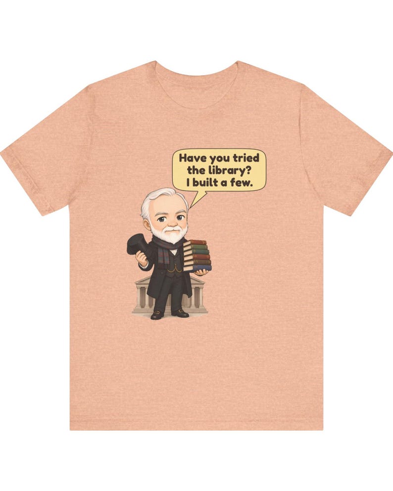 May include: A peach-colored t-shirt featuring a cartoon illustration of a bearded man in a suit holding a stack of books and a hat. A speech bubble above him contains the text: Have you tried the library? I built a few.
