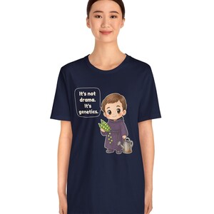 May include: A navy blue short-sleeve t-shirt featuring a cartoon illustration of a monk holding a pea pod and a watering can. The text in a speech bubble reads, "It's not drama. It's genetics." The design is centered on the front of the shirt.