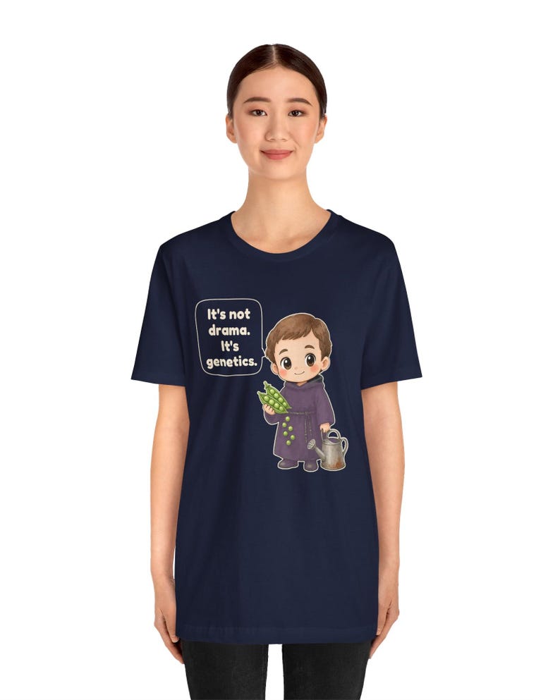 May include: A navy blue short-sleeve t-shirt featuring a cartoon illustration of a monk holding a pea pod and a watering can. The text in a speech bubble reads, "It's not drama. It's genetics." The design is centered on the front of the shirt.