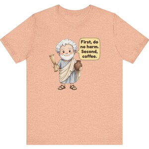 May include: A peach-colored t-shirt featuring a cartoon illustration of a white-haired man in a toga holding a scroll and a brown mug. A speech bubble next to the figure contains the text: First, do no harm. Second, coffee.