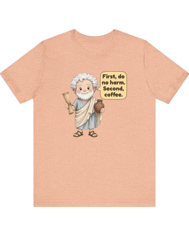 May include: A peach-colored t-shirt featuring a cartoon illustration of a white-haired man in a toga holding a scroll and a brown mug. A speech bubble next to the figure contains the text: First, do no harm. Second, coffee.