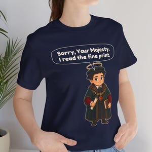 May include: A navy blue t-shirt featuring a graphic of a cartoon character wearing a dark robe, a gold chain, and a halo. A speech bubble above the character contains the text: Sorry, Your Majesty. I read the fine print. The shirt has short sleeves.