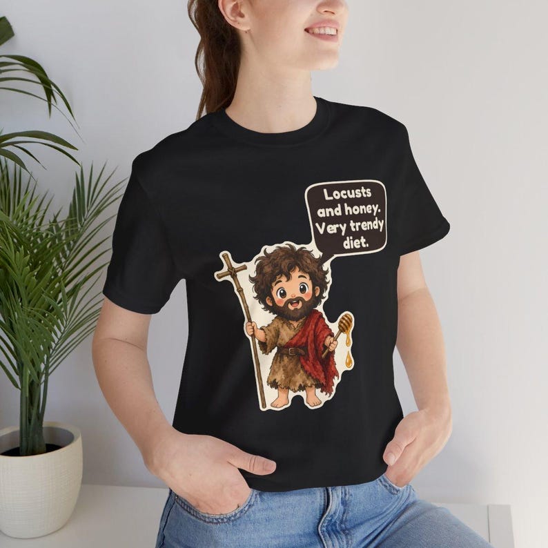 May include: A black t-shirt featuring a cartoon illustration of a bearded figure holding a wooden cross staff and a honey dipper. A speech bubble above the figure contains the text: Locusts and honey. Very trendy diet.