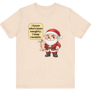 May include: A light beige t-shirt featuring a cartoon illustration of Santa Claus holding a scroll. A yellow speech bubble above Santa contains the text: I know who's been naughty. I keep receipts. Santa wears a red suit with white trim.