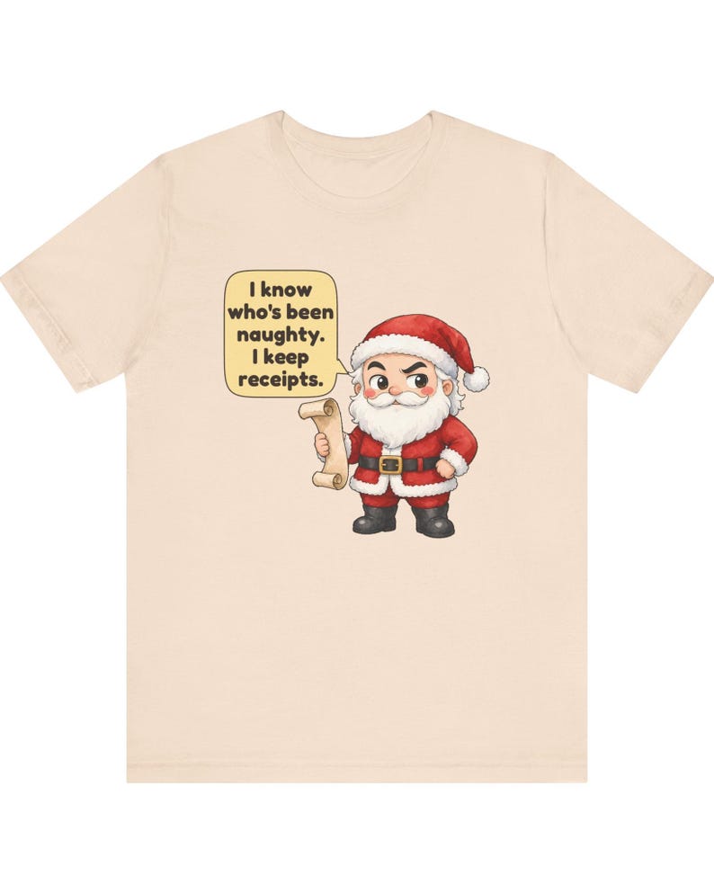 May include: A light beige t-shirt featuring a cartoon illustration of Santa Claus holding a scroll. A yellow speech bubble above Santa contains the text: I know who's been naughty. I keep receipts. Santa wears a red suit with white trim.