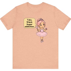 May include: A peach-colored short-sleeve t-shirt featuring a graphic of a cartoon ballerina with pink hair, a tiara, and a pink tutu. The character holds a star-tipped wand and a cupcake, with a speech bubble that reads: Tutu. Wand. Sugar addiction.