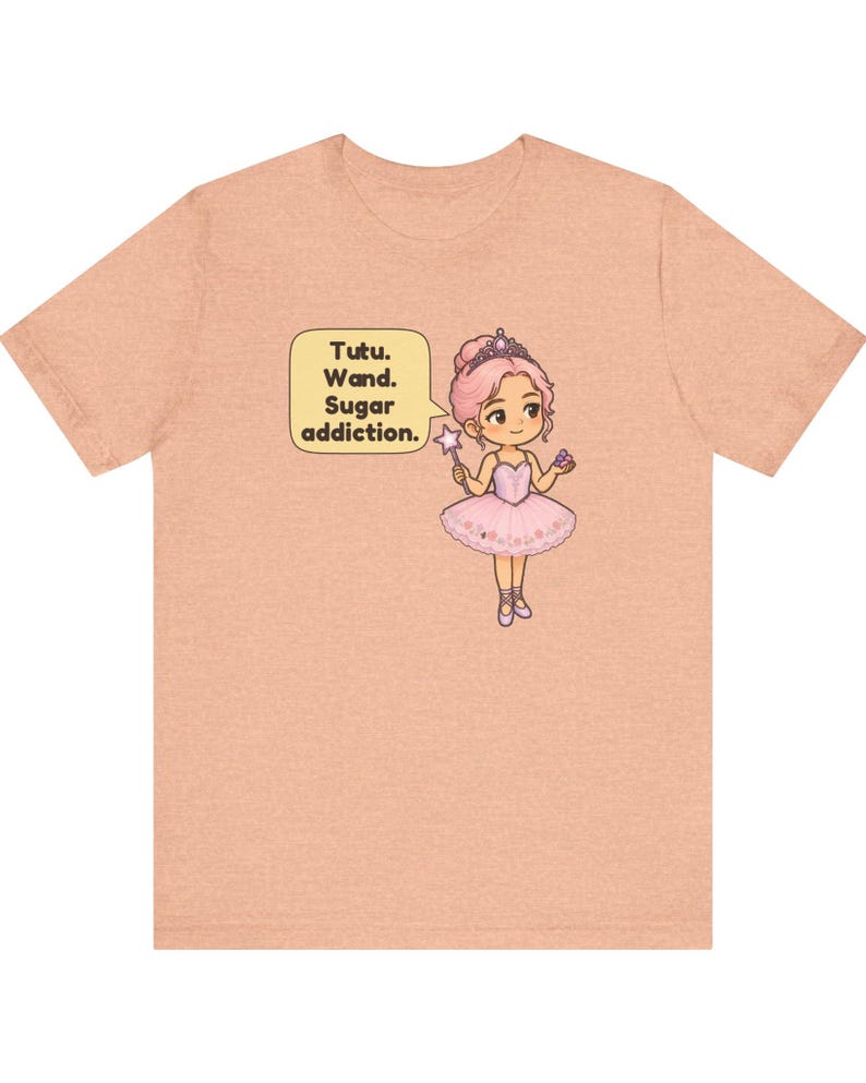 May include: A peach-colored short-sleeve t-shirt featuring a graphic of a cartoon ballerina with pink hair, a tiara, and a pink tutu. The character holds a star-tipped wand and a cupcake, with a speech bubble that reads: Tutu. Wand. Sugar addiction.