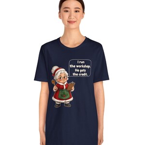 K&ouml;nnte beinhalten: Ein dunkelblaues Kurzarm-T-Shirt mit einer Cartoon-Illustration einer &auml;lteren Frau in einem roten Kleid und einer gr&uuml;nen Sch&uuml;rze, die einen Holzl&ouml;ffel und ein Klemmbrett h&auml;lt. Eine Sprechblase enth&auml;lt den Text: I run the workshop. He gets the credit.