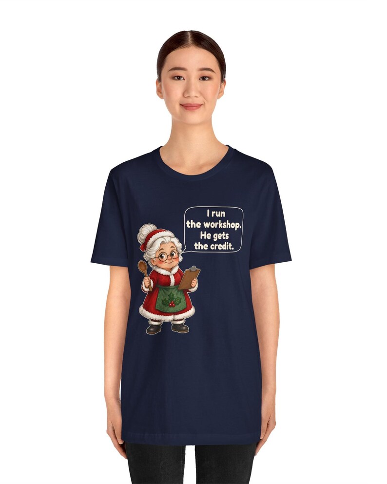 K&ouml;nnte beinhalten: Ein dunkelblaues Kurzarm-T-Shirt mit einer Cartoon-Illustration einer &auml;lteren Frau in einem roten Kleid und einer gr&uuml;nen Sch&uuml;rze, die einen Holzl&ouml;ffel und ein Klemmbrett h&auml;lt. Eine Sprechblase enth&auml;lt den Text: I run the workshop. He gets the credit.