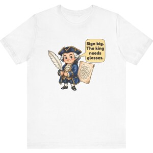 May include: A white t-shirt featuring a cartoon illustration of a historical figure in a blue coat and tricorn hat holding a quill and parchment. A speech bubble contains the text: Sign big. The king needs glasses.