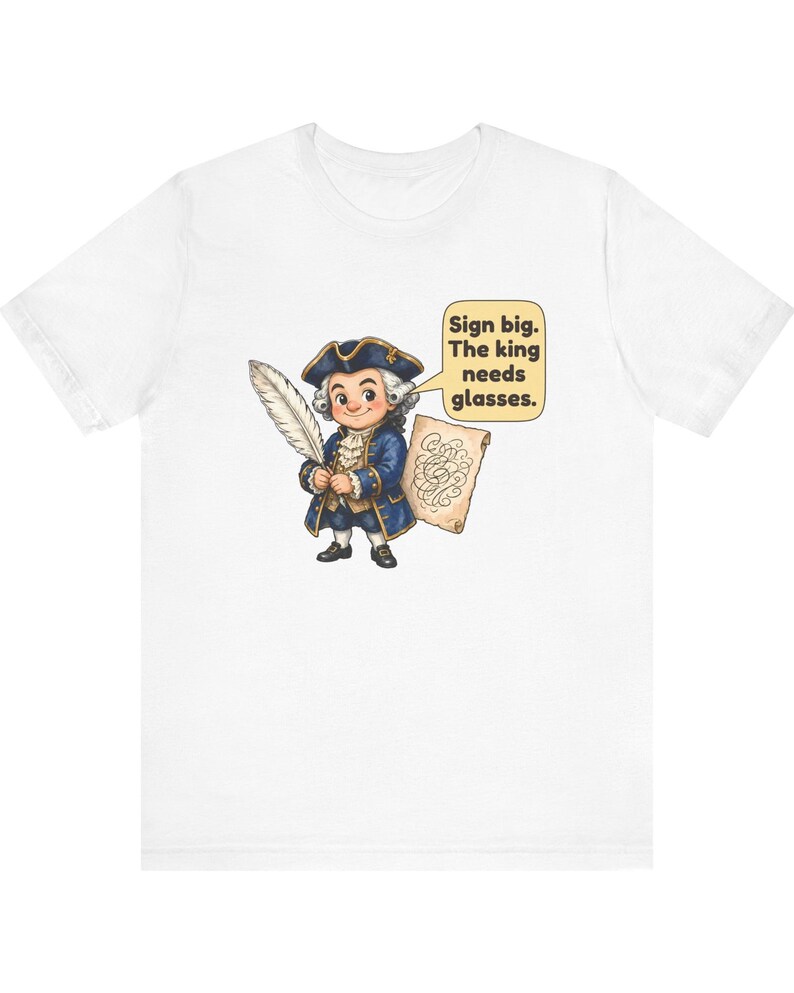 May include: A white t-shirt featuring a cartoon illustration of a historical figure in a blue coat and tricorn hat holding a quill and parchment. A speech bubble contains the text: Sign big. The king needs glasses.