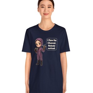 Puede incluir: Una camiseta azul marino con un personaje de dibujos animados con un traje de vuelo morado y botas oscuras, sosteniendo unas gafas. Un globo de texto contiene: I flew the Channel. Nobody noticed. El dise&ntilde;o est&aacute; centrado en el pecho.