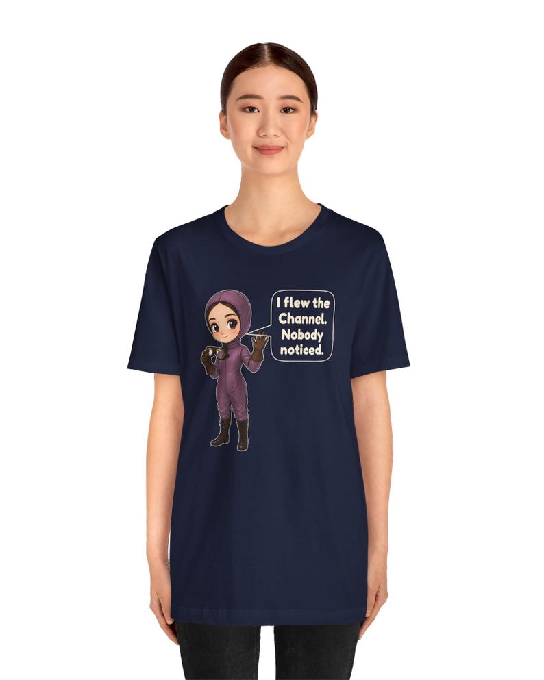 Puede incluir: Una camiseta azul marino con un personaje de dibujos animados con un traje de vuelo morado y botas oscuras, sosteniendo unas gafas. Un globo de texto contiene: I flew the Channel. Nobody noticed. El dise&ntilde;o est&aacute; centrado en el pecho.