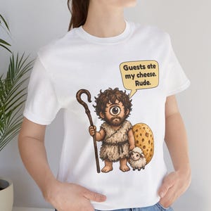 May include: A white t-shirt featuring a cartoon cyclops holding a wooden staff and standing next to a small sheep and a large piece of Swiss cheese. A speech bubble above the cyclops contains the text: Guests ate my cheese. Rude.