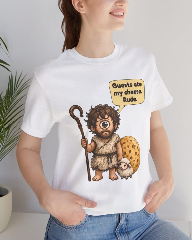 May include: A white t-shirt featuring a cartoon cyclops holding a wooden staff and standing next to a small sheep and a large piece of Swiss cheese. A speech bubble above the cyclops contains the text: Guests ate my cheese. Rude.