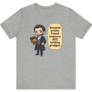 May include: A heather gray t-shirt featuring a cartoon illustration of a man in a dark coat holding a treasure chest, with a speech bubble containing the text: Escaped prison. Found treasure. Still holding grudges.