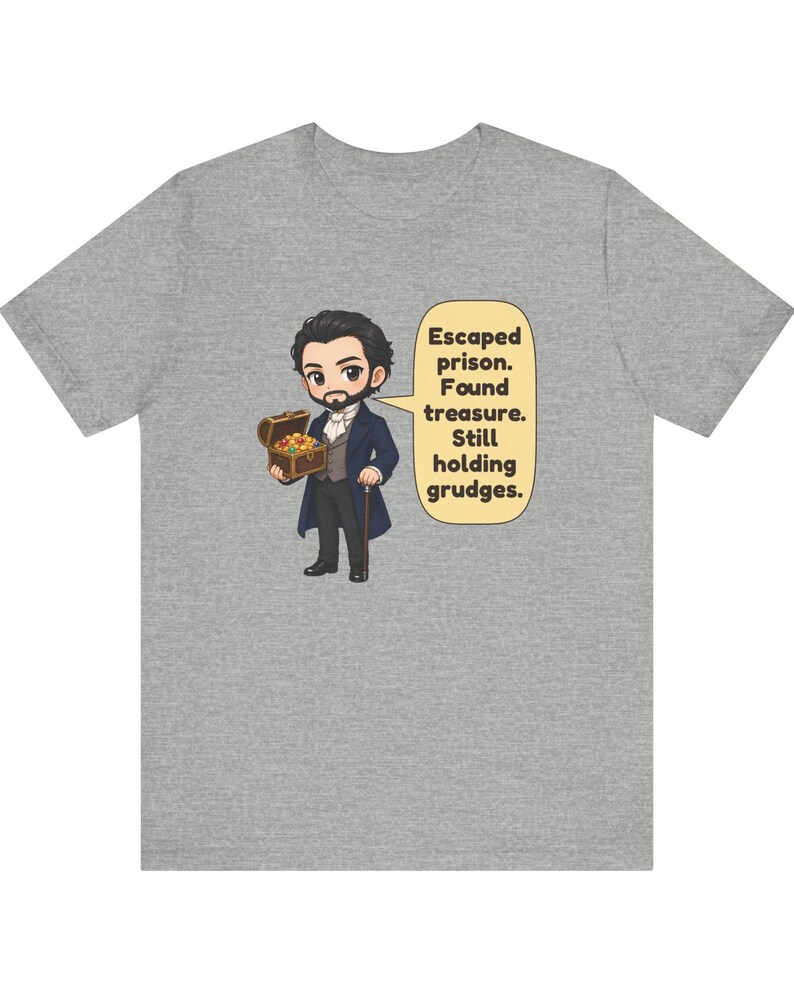 May include: A heather gray t-shirt featuring a cartoon illustration of a man in a dark coat holding a treasure chest, with a speech bubble containing the text: Escaped prison. Found treasure. Still holding grudges.