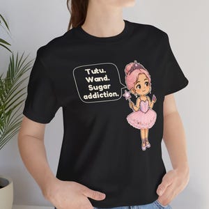 May include: A black t-shirt featuring a graphic of a cartoon ballerina with pink hair, a tiara, and a pink tutu, holding a star-shaped wand. A speech bubble next to the character contains the text: Tutu. Wand. Sugar addiction.