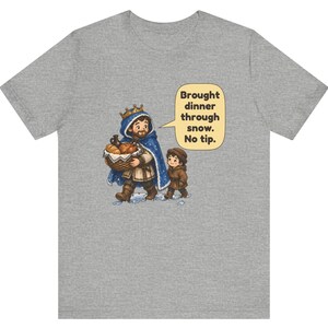 May include: A heather gray t-shirt featuring a cartoon illustration of a king in a blue cloak carrying a basket of bread and a child walking in snow. A speech bubble contains the text: Brought dinner through snow. No tip.