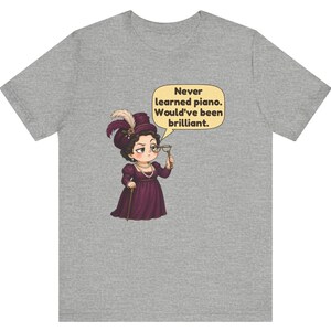May include: A heather gray t-shirt featuring a cartoon illustration of a person in a dark purple Victorian-style dress and hat, holding a monocle. A speech bubble contains the text: Never learned piano. Would've been brilliant.