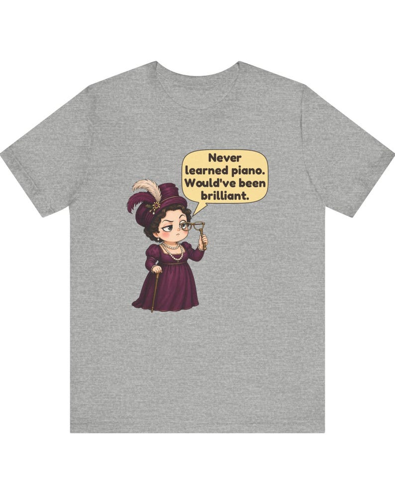 May include: A heather gray t-shirt featuring a cartoon illustration of a person in a dark purple Victorian-style dress and hat, holding a monocle. A speech bubble contains the text: Never learned piano. Would've been brilliant.