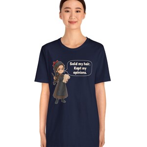 May include: A navy blue short-sleeve t-shirt featuring a cartoon illustration of a character with a red hair bow, wearing a brown dress and apron, holding a paper. A speech bubble contains the text: Sold my hair. Kept my opinions.