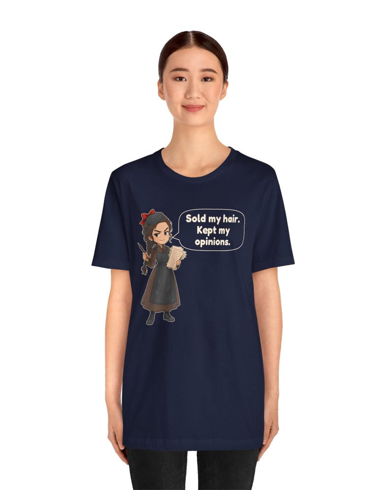 May include: A navy blue short-sleeve t-shirt featuring a cartoon illustration of a character with a red hair bow, wearing a brown dress and apron, holding a paper. A speech bubble contains the text: Sold my hair. Kept my opinions.