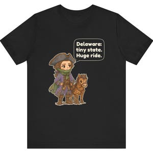 May include: A black t-shirt featuring a cartoon illustration of a person in a tricorn hat and colonial-style clothing standing next to a small brown horse. A speech bubble above them contains the text: Delaware: tiny state. Huge ride.