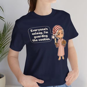Op de afbeelding: Een marineblauw t-shirt met een afbeelding van een personage in een rood-wit gestreepte pyjama die een kaars en een bord met koekjes vasthoudt. Een tekstballon bevat de tekst: Everyone's asleep. I'm guarding the cookies.